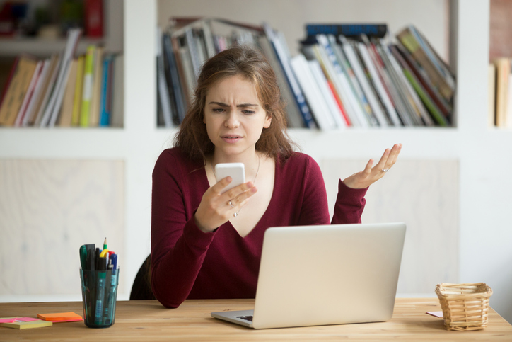 woman looking at phone frustrated while sitting behind a laptop at a desk